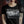 A woman wearing a black short-sleeve graphic T-shirt with a large black-and-white 'Brodequin' band print on the front and a circular white emblem on the sleeve, photographed against a dark brick wall background.