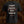 A black short-sleeve Liturgy band t-shirt shown from the back, featuring a gothic-style white and red graphic print with the text 'everyone has gone to the divination' and occult geometric symbols. The shirt is displayed against a dark, textured brown background.
