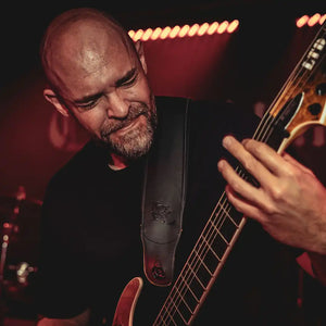 A close-up of Brodequin guitarist Mike Bailey playing guitar on a stage, wearing a black shirt and a black leather guitar strap with a debossed logo.
