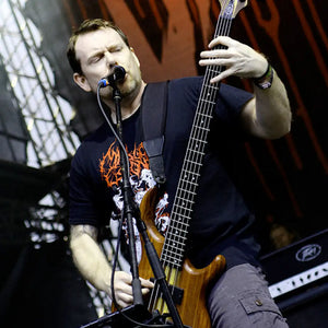 Brodequin bassist and vocalist Jamie Bailey performing live on stage, playing a dark-colored five-string bass guitar and growling into a microphone under dramatic blue stage lighting.