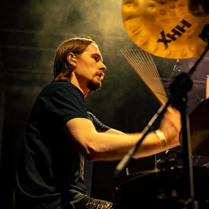 Brodequin drummer Brennan Shackelford in a black t-shirt playing on stage under dramatic warm lighting, with a gold cymbal visible in the background.